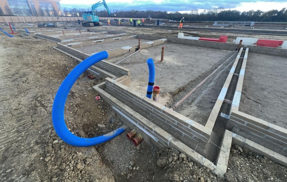 Construction site with foundation work, blue drainage pipes, and workers in the background.