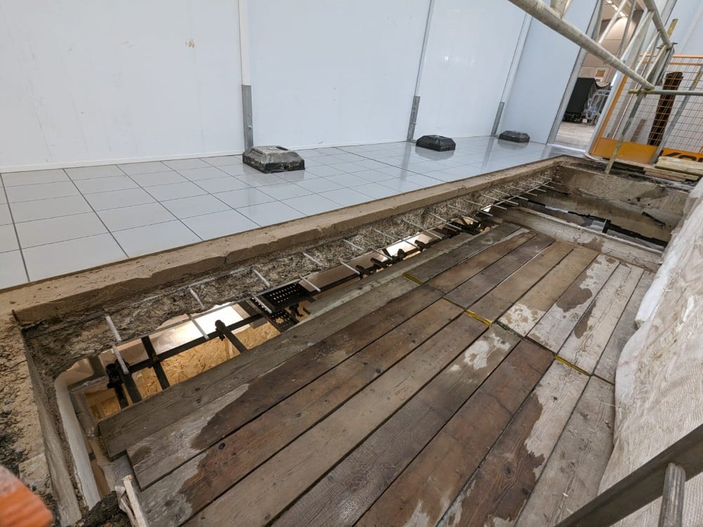 Large rectangular hole in the floor exposing wooden boards and construction materials in an indoor area.