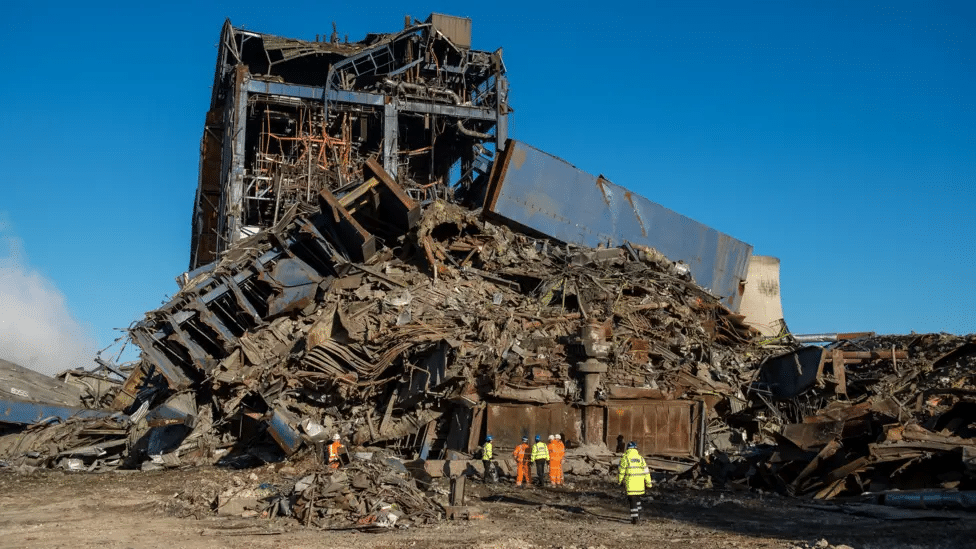 Collapsed power station building with rubble and workers in high-visibility suits in the foreground.