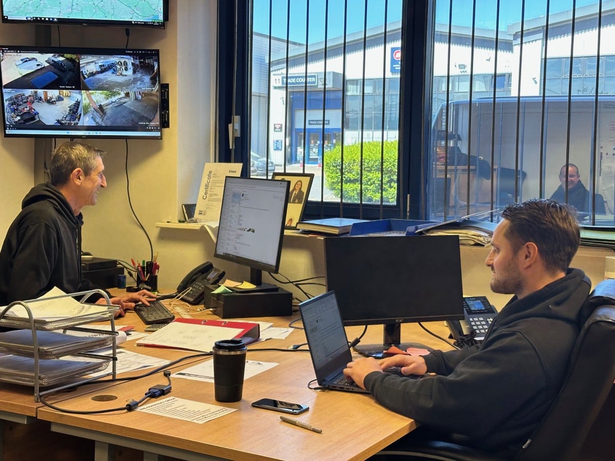 Two men working at desks with computers and security monitors in a bright office.