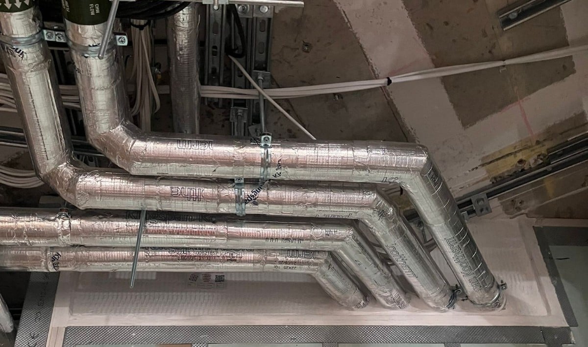 Insulated metal pipes run along a ceiling, fitted with fire sleeves for protection.