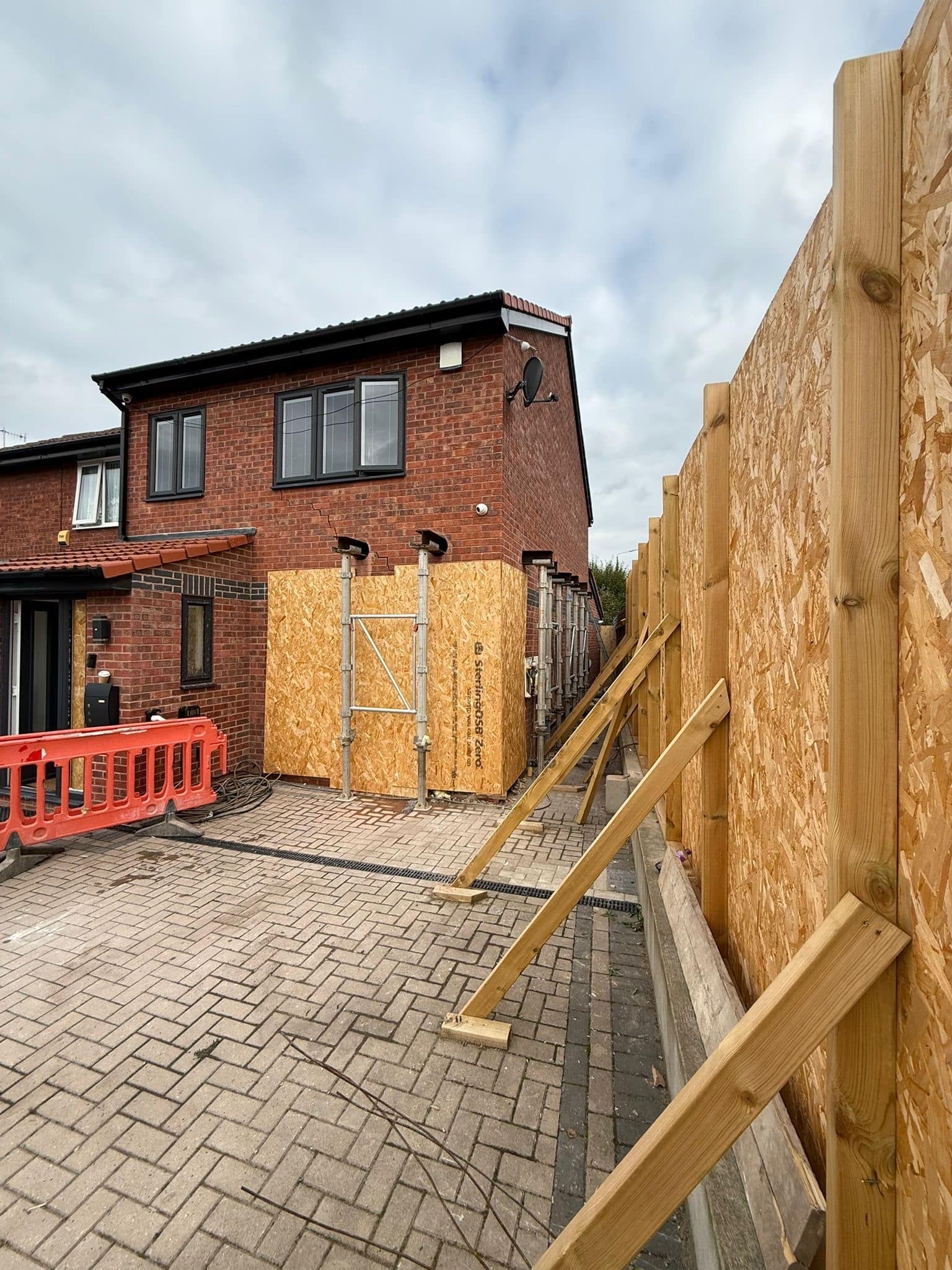 Wooden temporary works and barriers set up outside a brick house under cloudy skies.