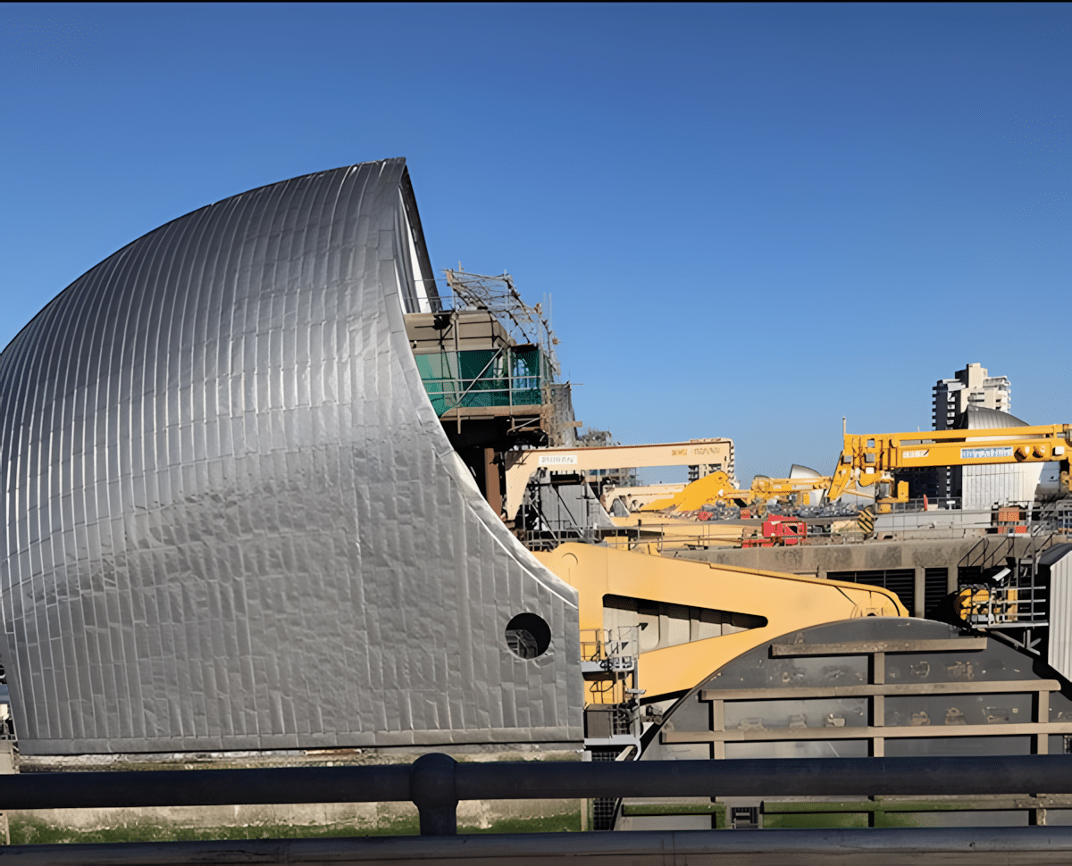 The Thames Barrier with construction equipment and buildings in the background under a clear sky.