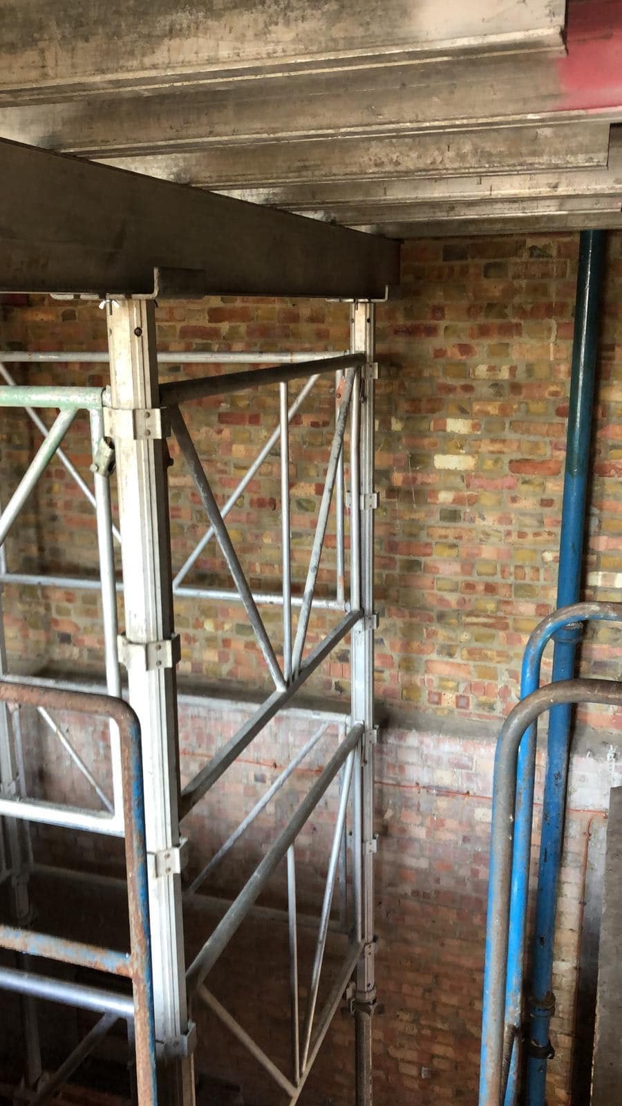 Metal scaffolding set up next to a brick wall with blue pipes in an indoor construction area.