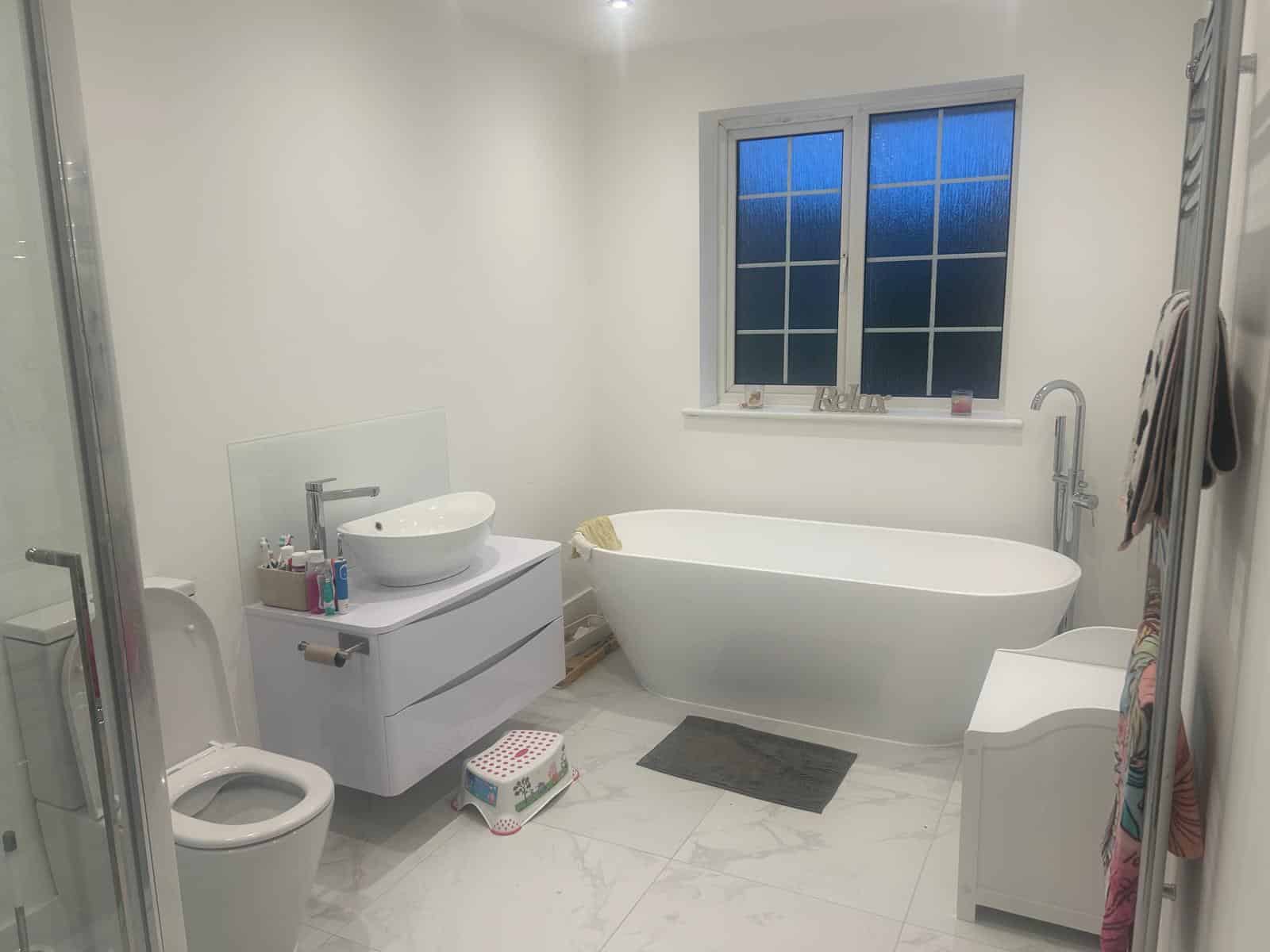 Modern bathroom with a bathtub, sink, toilet, and window, featuring white walls and floor.