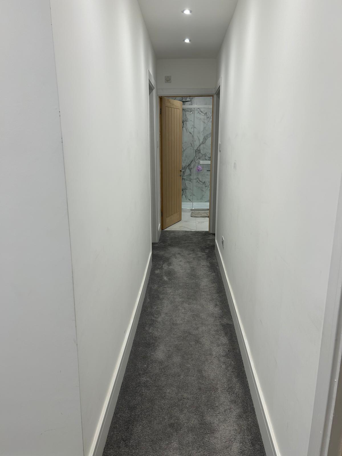 A narrow hallway with grey carpet leads to an open door and a room with a marble wall.