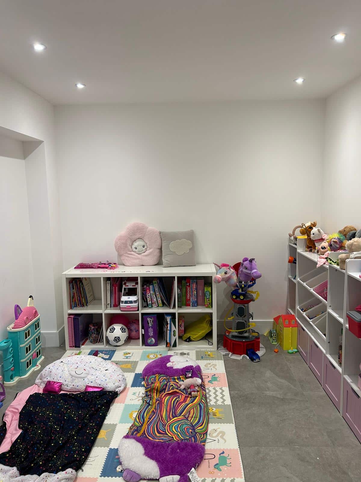Children’s playroom with shelves of toys and books, colorful bedding, and stuffed animals on the floor.