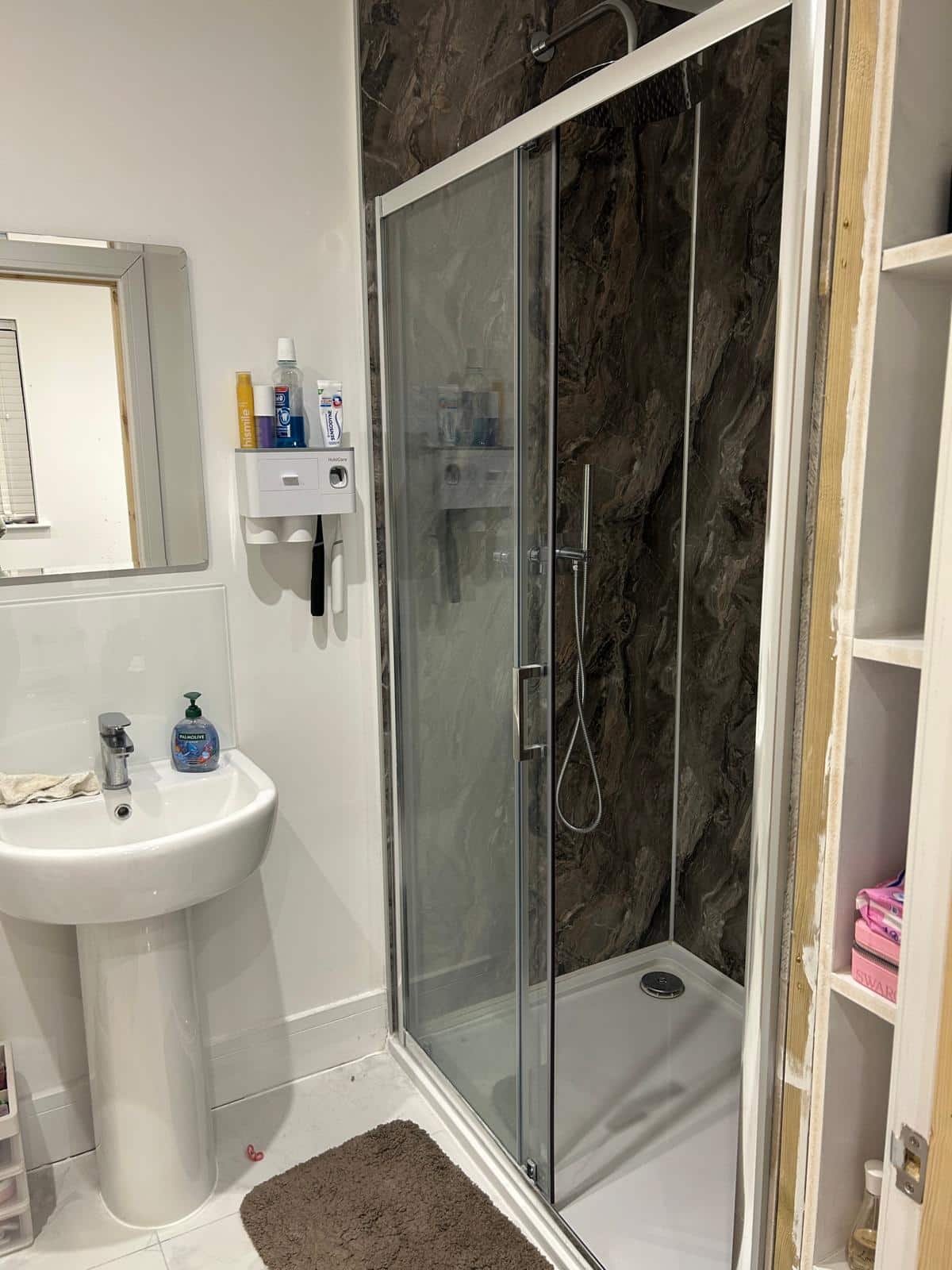 A small bathroom with a glass shower, pedestal sink, mirror, and shelves with towels and toiletries.