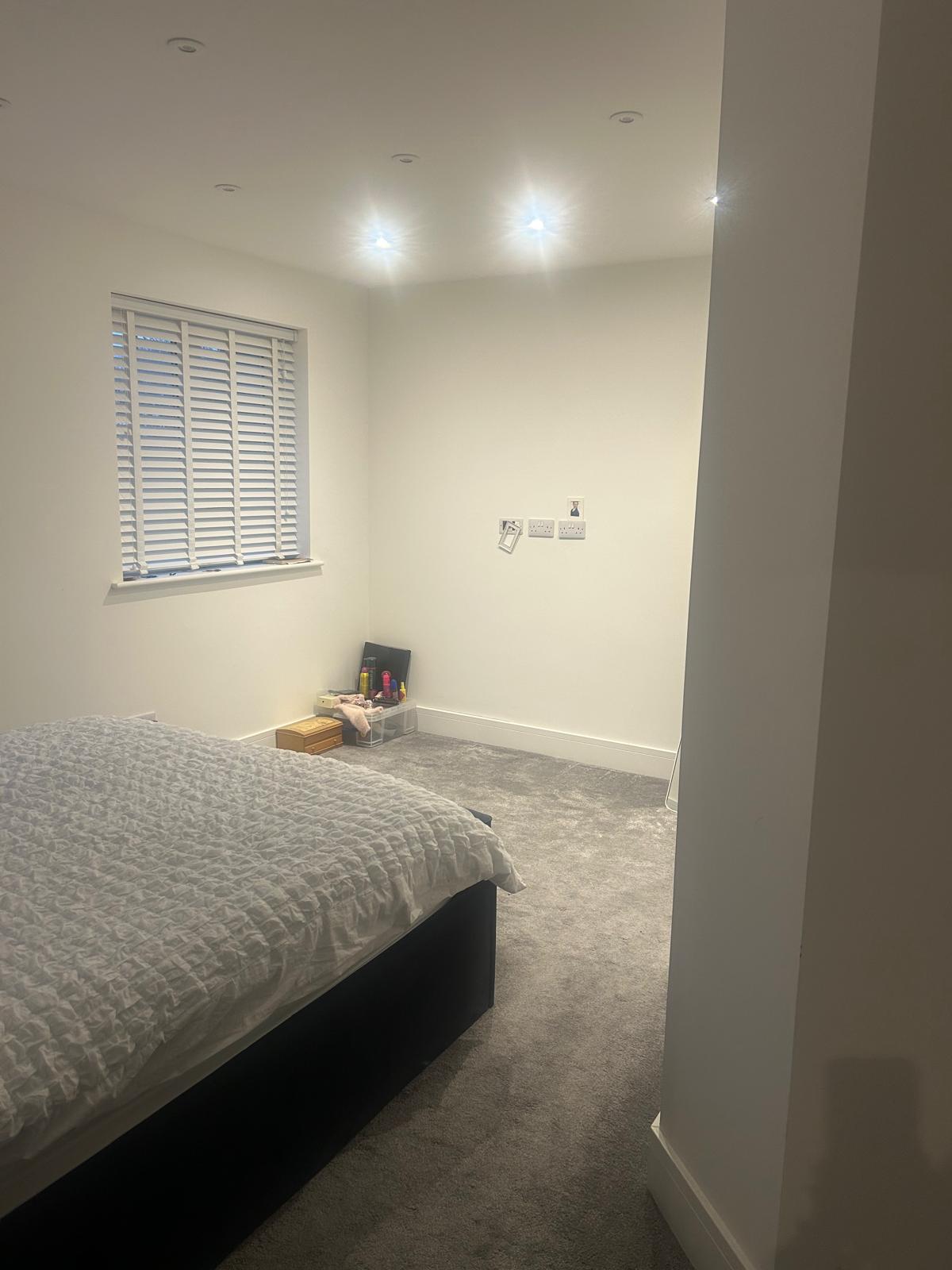 A simple bedroom with a bed, window blinds, and a few items on the carpeted floor.