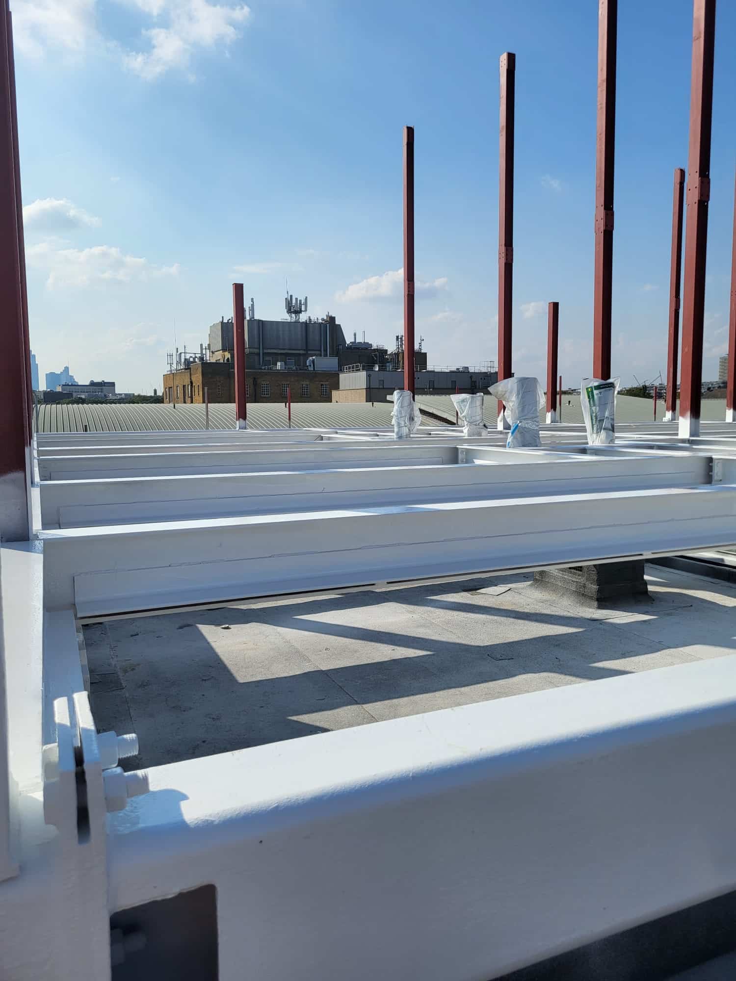 firestopping12 White metal beams and vertical red rods on a rooftop construction site under a blue sky.