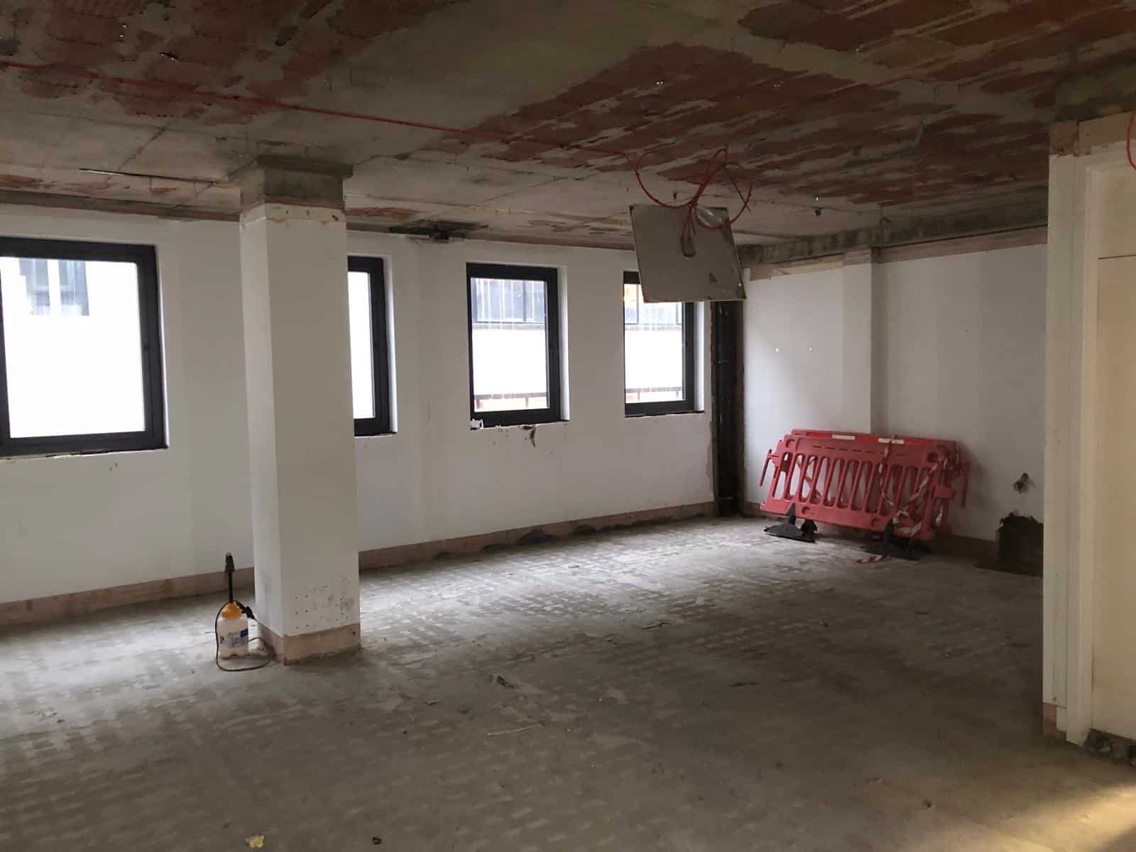 jermyn street3 Empty room under renovation with three windows, exposed ceiling, and construction materials in the corner.