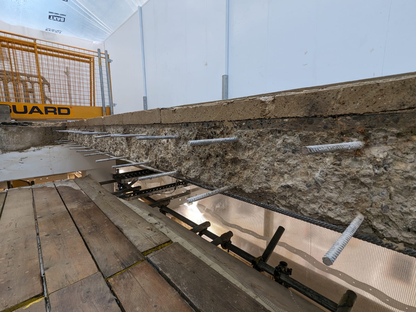Concrete structure under repair with exposed steel rods and wooden planks nearby.