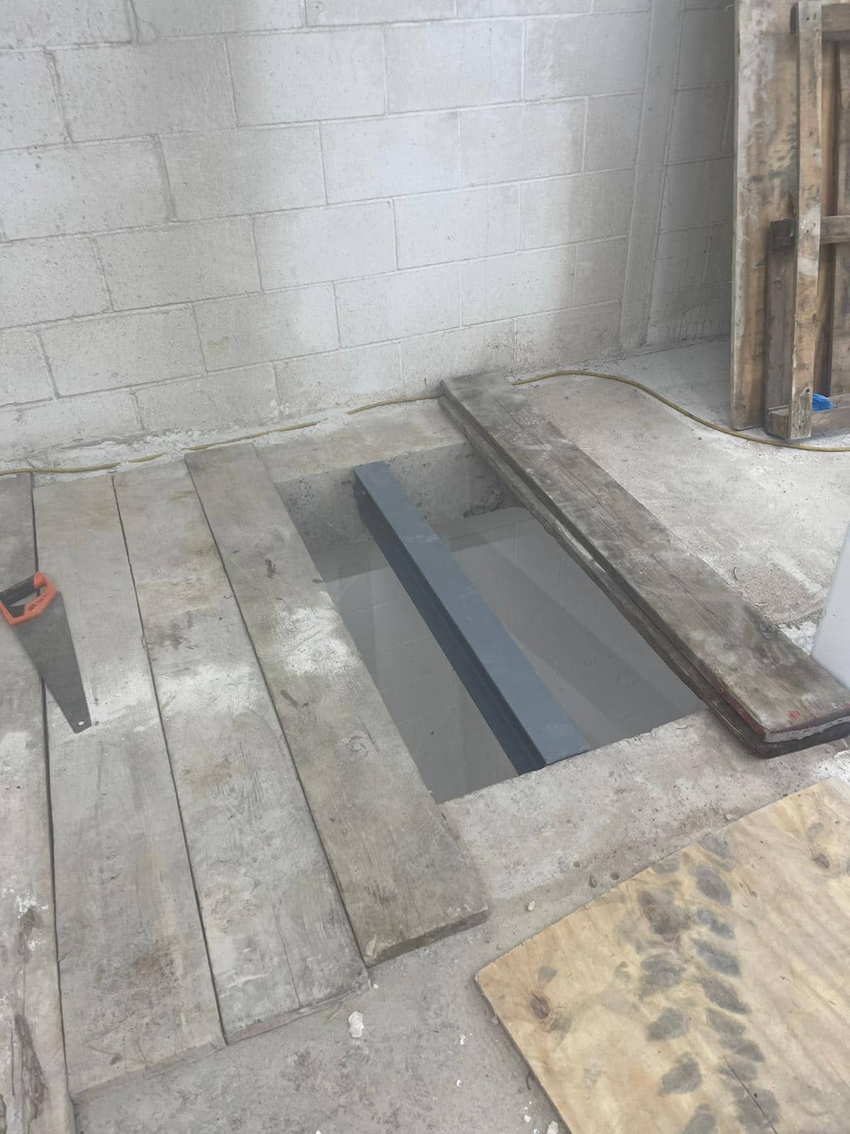 steels07 Wooden planks covering a rectangular hole in a concrete floor by a cinder block wall.